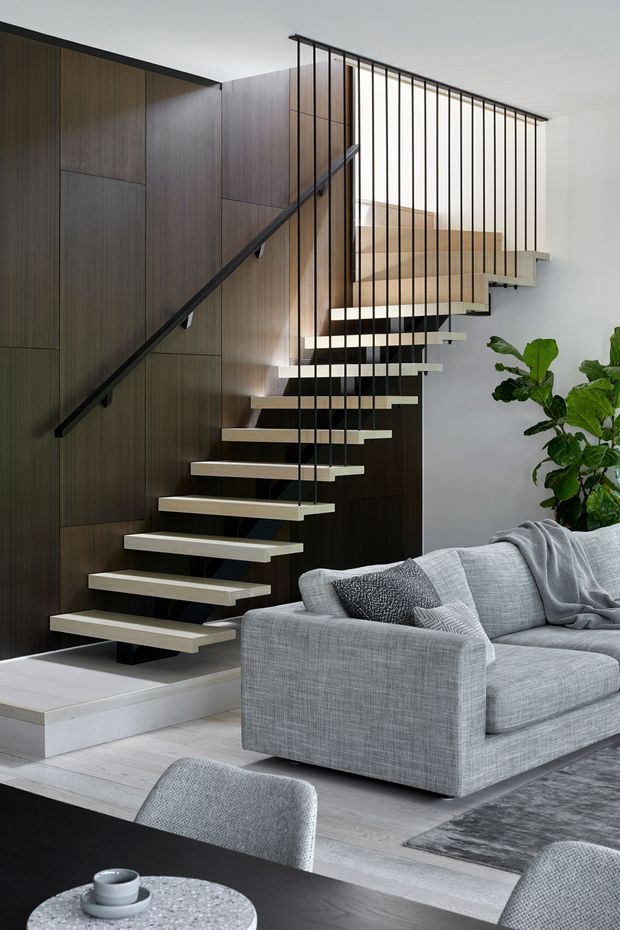 Contemporary open-plan living area featuring a sculptural floating staircase with pale timber treads, dark vertical metal balustrade rods, and dark timber wall panelling. A large grey sectional sofa, textured rug, dining table and upholstered chairs occupy the foreground, while a leafy indoor plant adds greenery. The space is bright, refined and minimalist in composition.