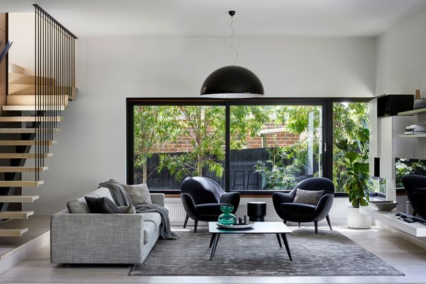 A refined contemporary living room with a grey sectional sofa, two dark lounge chairs, a low black coffee table, and a large grey area rug. Full-width black-framed glazing opens views to a leafy courtyard, while a floating timber staircase with a slender black vertical balustrade defines the left side. White walls, built-in white media joinery, indoor plants, and a large black dome pendant complete the clean, modern interior.