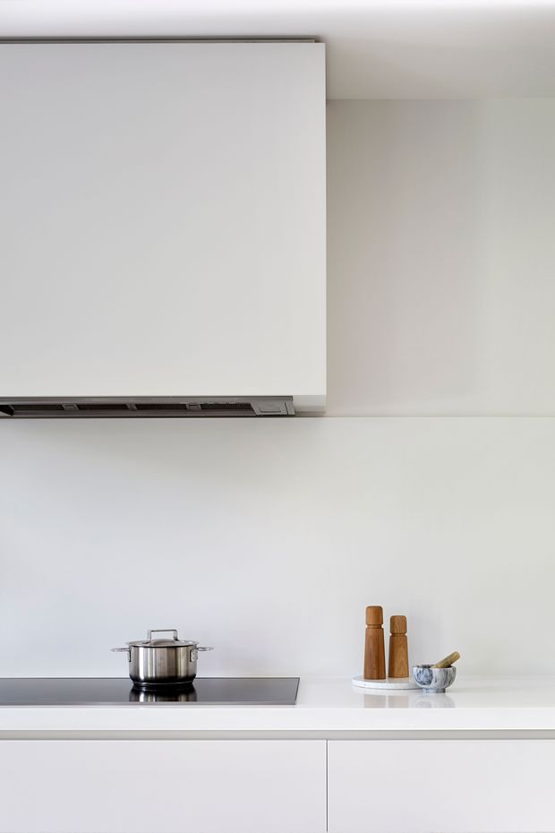 Close-up of a minimalist contemporary kitchen featuring a white induction cooktop with a stainless-steel pot, a large integrated white rangehood, white splashback and benchtop, and streamlined handleless white cabinetry. Small wooden grinders and a mortar and pestle provide the only visible accents.