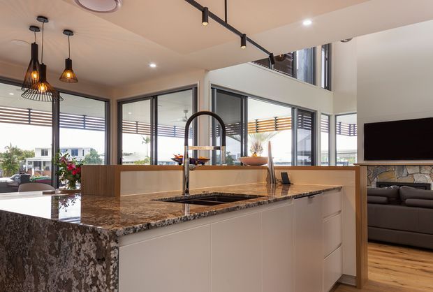 Bright contemporary open-plan kitchen with a large waterfall-edge stone island, integrated sink and tall gooseneck faucet. White flat-panel cabinetry, dark veined stone surfaces, pendant lighting, black-framed glazing, timber flooring and an adjacent living area with a television are visible. The space opens toward an outdoor terrace and appears to include a high void or upper-level glazing.