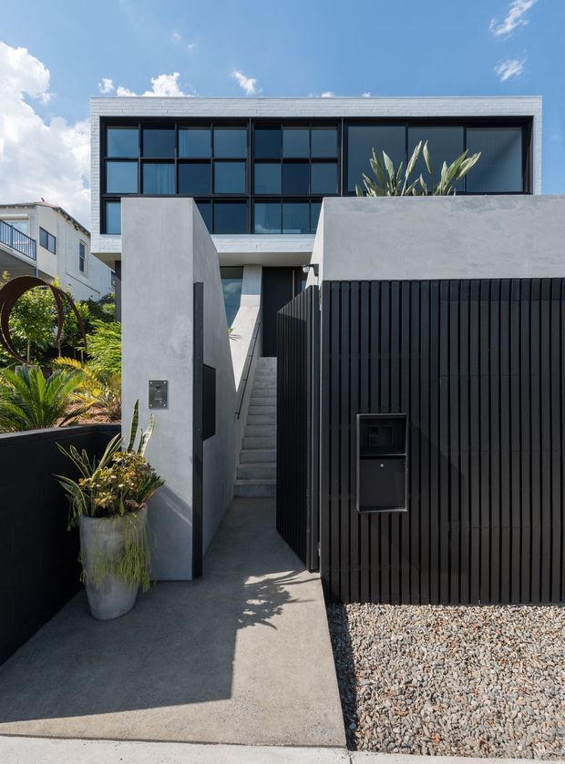 Front exterior view of a contemporary multi-storey residence. The entry is approached via a narrow concrete walkway and stair, framed by tall smooth-rendered walls and a tall black vertical-slat gate with integrated letterbox. The upper facade features extensive dark-framed grid glazing, while gravel and planted areas soften the minimalist street frontage.