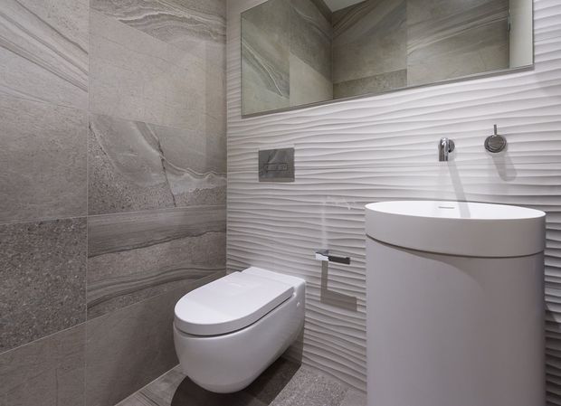 Contemporary compact bathroom featuring a wall-hung toilet, sculptural freestanding basin, wall-mounted chrome tapware, large-format grey stone-look wall and floor tiles, and a white three-dimensional textured feature wall beneath a mirrored cabinet.