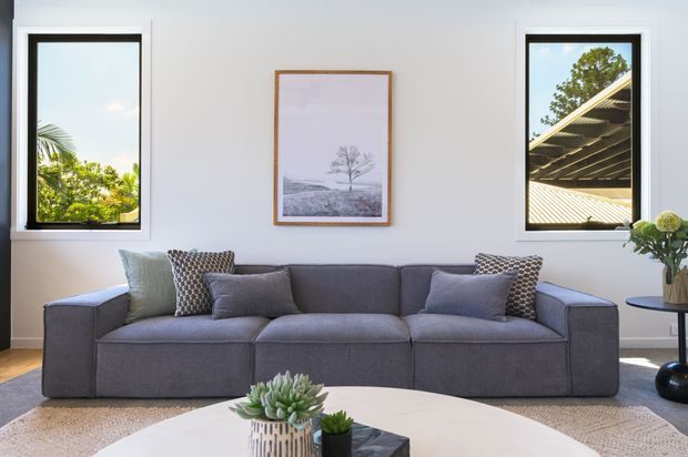 Bright, contemporary living room with a large charcoal-grey modular sofa, neutral cushions, a round light coffee table, indoor plants, and framed artwork. White walls and black-framed windows bring in abundant natural light, with greenery and a roof structure visible outside.