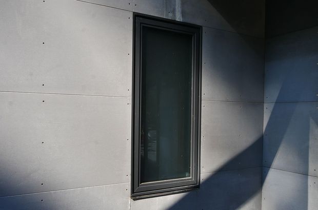 Close-up exterior view of a narrow dark-framed window set into large smooth grey wall panels. Strong directional sunlight creates angular shadows across the minimalist facade, with visible panel joints and fasteners.