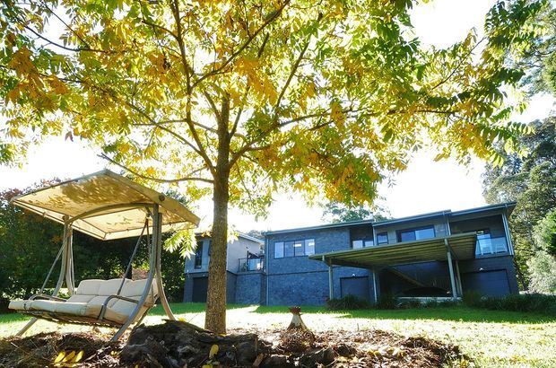 Exterior view of a contemporary two-storey dark-clad house set in a leafy landscaped garden. The home has a low-pitched roof, broad horizontal proportions, large rectangular windows, and a covered projecting terrace or balcony. A freestanding outdoor swing seat is visible in the foreground beneath a mature deciduous tree.