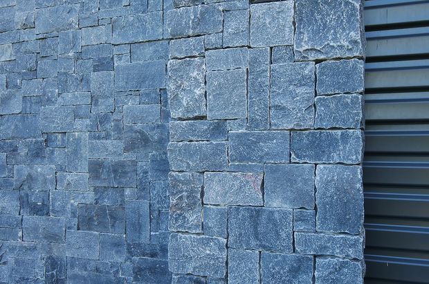 Close-up of a blue-grey natural stone masonry wall with irregular rectangular blocks and pronounced rough, textured faces. A dark horizontal metal-clad or louvred surface is visible along the right edge, suggesting an exterior architectural facade detail. No interior room or furnishings are visible.