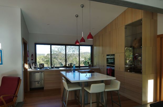 A contemporary kitchen and dining area with extensive warm timber cabinetry, a central island with a pale stone benchtop, integrated appliances, and a glass-front display cabinet. Three red pendant lights hang above the island, while large windows provide views to greenery outside. A small dining setting is positioned at the island, with dark timber flooring and a bright, open atmosphere.