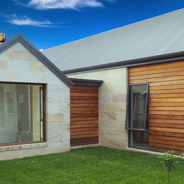 Exterior view of a low-profile residential building with a pitched corrugated metal roof, pale rectangular stone cladding, and warm horizontal timber wall cladding. Dark-framed glazing and a small manicured lawn define the courtyard-facing facade.