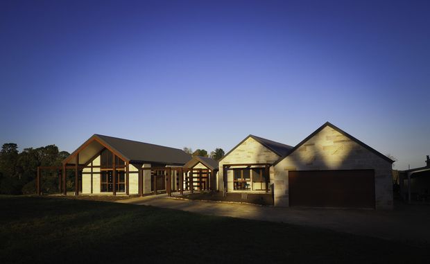 Single-storey residential house photographed at dusk, with multiple connected pitched-gable roof forms, timber-framed glazed sections, pale masonry or rendered walls, and a dark double garage door. The home sits within a landscaped suburban or semi-rural setting.