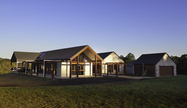 A single-storey rural residential complex photographed at dusk, with several connected gabled forms. The central building has a strong timber post-and-beam structure, extensive glazing, dark metal pitched roofs, and warm timber framing. Separate smaller gabled wings or outbuildings sit to either side, with open grass and trees surrounding the property.