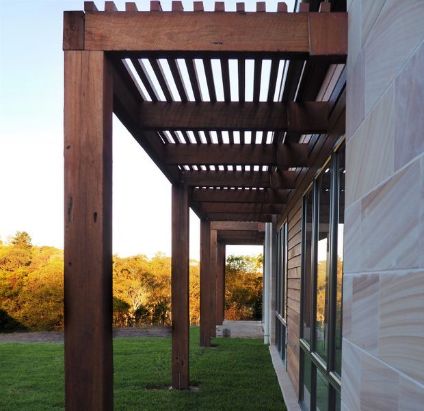 Exterior view along a covered timber pergola or colonnade attached to a contemporary building. The structure features substantial dark-stained timber posts and beams with regularly spaced overhead slats, beside full-height glazed doors or windows and horizontal wall cladding. A lawn and landscaped vegetation are visible beyond.