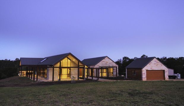 Twilight exterior view of a contemporary rural residential complex comprising a prominent glazed gable-roof house with warm interior lighting, flanked by additional gabled buildings and a detached garage. The buildings use dark metal roofing, light masonry walls, extensive window glazing, and timber-toned garage doors, set on an open grassy site.