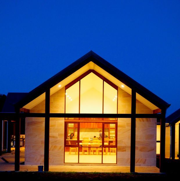 Front-facing dusk view of a modern gabled building with a strongly illuminated glazed entrance facade. Dark structural framing contrasts with warm timber-toned interior lighting and pale masonry wall panels; a dining or communal space is visible through the central glazing.
