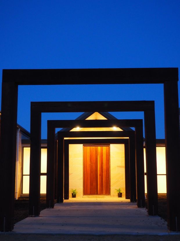 Symmetrical dusk view of a modern residential entrance framed by a sequence of large black rectangular portal structures. A concrete path leads to a warm timber pivot door set within a pale tiled facade, with concealed and recessed lighting illuminating the entry.