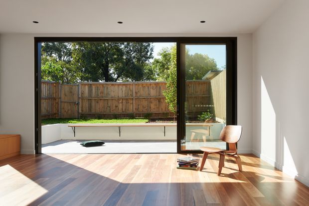 Bright, minimalist interior room opening through wide dark-framed glazed doors to a private landscaped courtyard. The space has warm timber flooring, white walls and ceiling, recessed downlights, and a mid-century-style wooden lounge chair. A built-in low outdoor planter or bench is visible beyond the glazing, with strong sunlight casting angular shadows across the floor.
