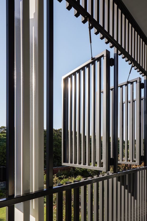 Close-up architectural view of a contemporary exterior screen or balustrade system, featuring repeated vertical metal slats in black and light grey, suspended or projecting panels, and strong geometric lines. Sunlight creates sharp highlights and shadows, with landscaped greenery visible beyond. The image focuses on the building facade and its screening/railing details against a clear blue sky.