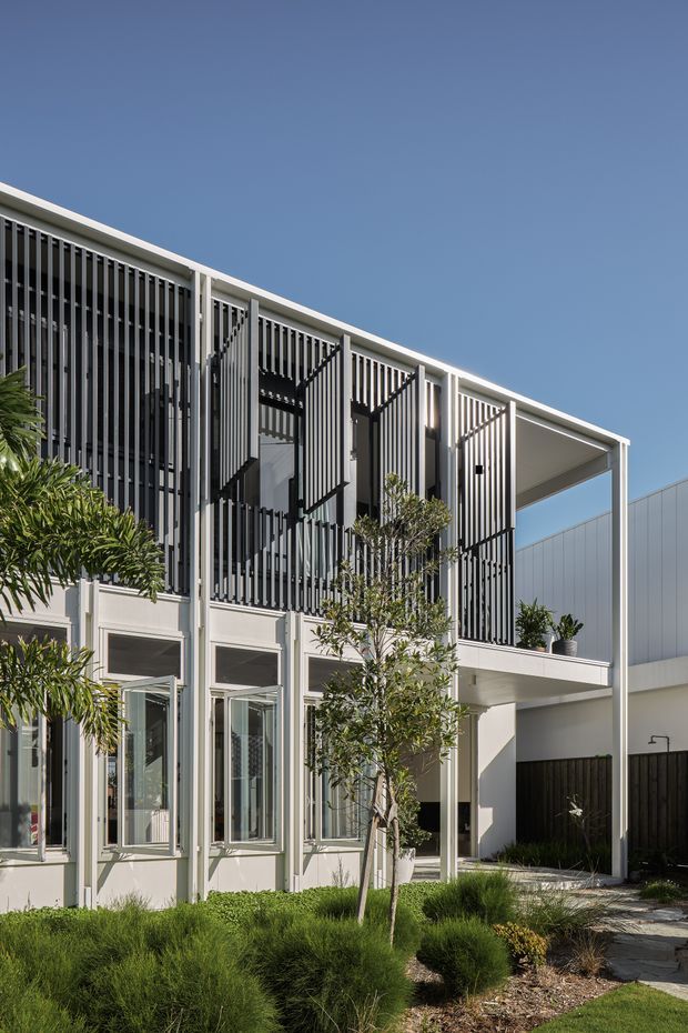 A modern two-storey residential building viewed from the landscaped front garden. The pale rendered facade has extensive dark vertical screening, projecting upper-level balcony or terrace areas, slim white structural posts, large glazed openings, and a simple flat roofline. Native-style grasses and shrubs soften the contemporary architecture, with a clear blue sky behind.