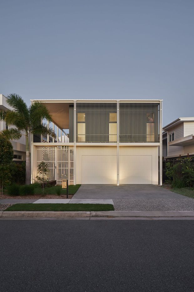 Street-facing view of a contemporary two-storey residential house with a prominent upper-level screened facade of closely spaced vertical slats. The pale rendered or clad structure includes a large enclosed garage at ground level, a concrete driveway, small landscaped front garden, and palm planting. The home has a flat or very low-pitched roofline and strong open-frame geometry, photographed at dusk from across the street.