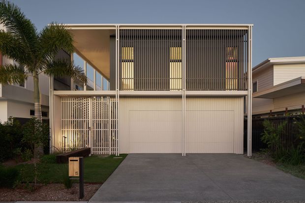 Street-facing view of a contemporary multi-level residence with a broad double garage, white vertical screening and balustrades, upper-level glazing, and a simple flat roofline. A concrete driveway, landscaped front garden, and tropical planting frame the facade.