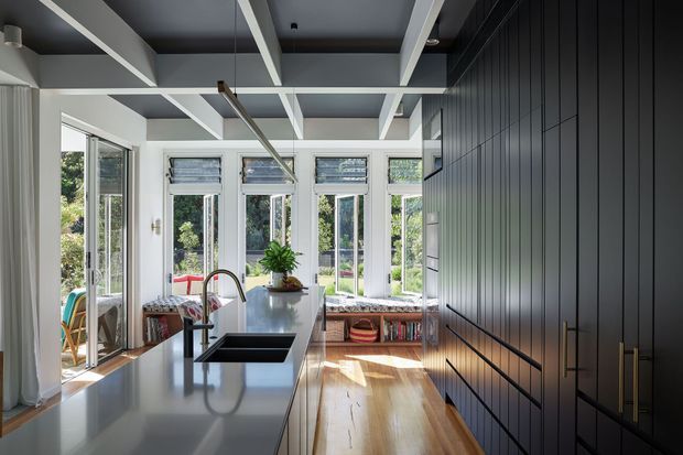 A bright contemporary kitchen viewed toward a wall of tall windows and glazed doors overlooking greenery. The room features a long pale island with an undermount sink and brushed metal gooseneck tap, white cabinetry, dark vertical-panel cabinetry along the right wall, integrated appliances, timber flooring, and a white-painted exposed beam ceiling with linear pendant lighting. A built-in window seat with patterned cushions is visible at the far end.