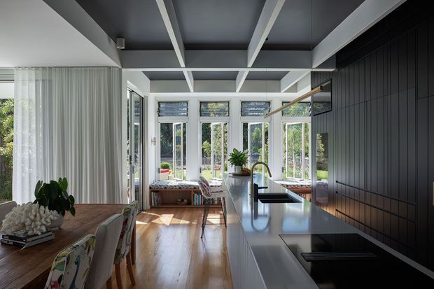 A bright, open-plan kitchen and dining area with extensive garden-facing glazing. The kitchen features a long island with a dark reflective benchtop, integrated black cooktop, stainless-steel tap and sink, and dark vertical wall panelling. White exposed ceiling beams contrast with the dark ceiling panels, while timber flooring and a timber dining table add warmth. Built-in window seating and indoor plants overlook the landscaped garden.