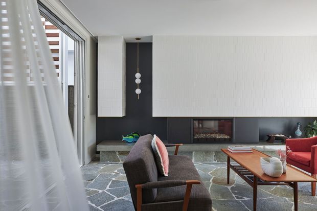A refined contemporary living room viewed through sheer curtains, featuring a dark fireplace wall with a recessed firebox, white vertical wall panelling, a grey stone slab floor, and expansive glazing. Mid-century-inspired lounge chairs and a timber coffee table furnish the room, with pendant lighting and muted decorative accents.