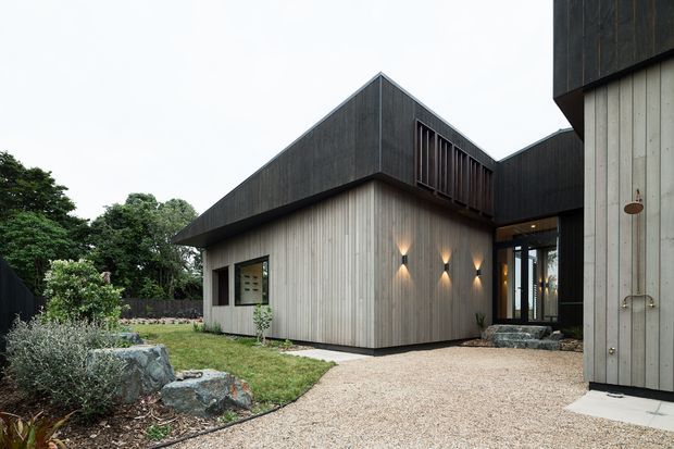 Exterior view of a contemporary single-storey residential building composed of interlocking gabled volumes. The facade combines dark vertical timber cladding on the upper forms with pale vertical timber boards below, illuminated by wall sconces. A gravel pathway, native-style landscaping, large rocks, and glazed entry areas are visible.