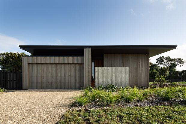 A modern single-storey house exterior with a broad low-pitched roof, vertical timber-clad walls, and a partially concealed recessed entry. The facade is viewed from a landscaped front approach, with a light aggregate driveway and ornamental grasses in the foreground.