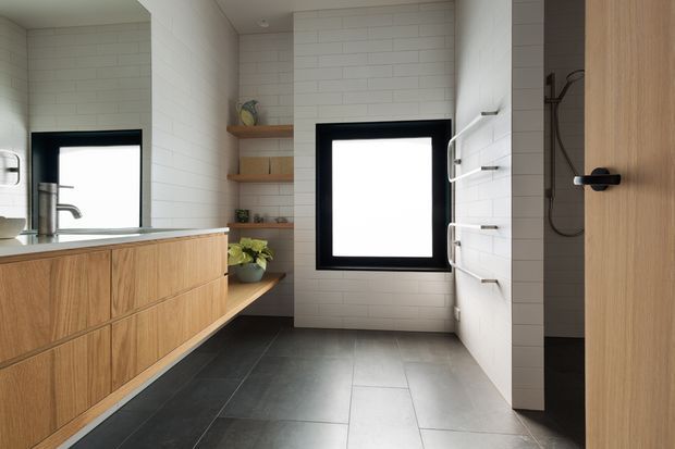 A bright contemporary bathroom with a floating timber vanity, integrated basin and chrome tap beneath a large mirror. White rectangular wall tiles, dark grey floor tiles, open timber shelving with plants, and a black-framed window create a clean, warm-modern scheme. A glass-enclosed shower with matte black fittings is visible at the right.