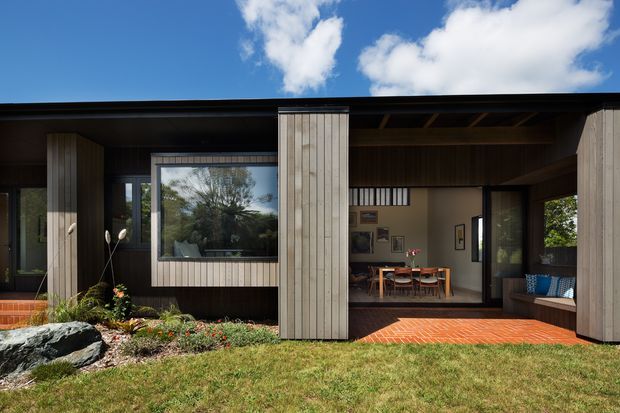 Single-storey modern home exterior with dark timber cladding, broad flat roof overhangs, and floor-to-ceiling glazing. A central timber-clad wall divides glazed living spaces, with a brick-paved terrace and built-in outdoor bench visible in the open recessed area. The landscaped foreground includes lawn, rocks, and planting, while an interior dining area is visible through the glazing.