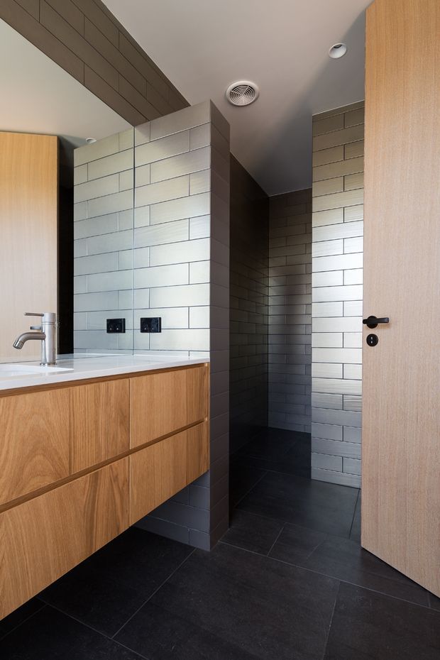 Contemporary bathroom interior featuring a floating timber vanity with integrated white basin and chrome tapware, a large mirrored wall, pale rectangular tiled walls, and dark charcoal floor tiles. A tiled shower or wet-area zone is visible beyond the partial-height tiled partition, with a light timber door and black hardware.