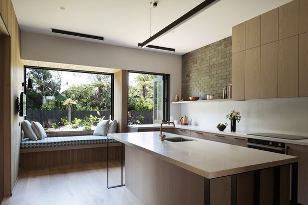 A bright, contemporary kitchen with a large white island and integrated cooktop, warm timber cabinetry, a pale brick feature wall, and broad windows overlooking a planted garden. A built-in window seat with cushions occupies the rear corner, while slim black linear lighting and dark window frames provide contrast.