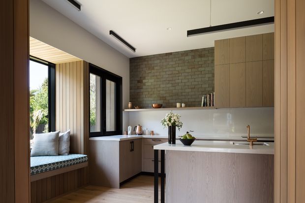 A warm contemporary kitchen with a light timber island, pale cabinetry, white benchtops, and a full-height exposed brick feature wall. The room includes a built-in window seat beside large black-framed windows, vertical timber wall panelling, sleek linear pendant and ceiling lighting, and views to greenery outside.