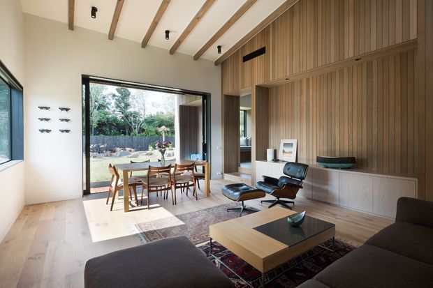 A bright, open-plan living and dining room with full-height glazing and an open doorway to a landscaped garden. The room features a timber-lined feature wall with integrated cabinetry and shelving, exposed timber ceiling beams, pale timber flooring, a mid-century lounge chair and ottoman, a large dark sectional sofa, a timber coffee table, and a dining table with wooden chairs. The interior has a warm, refined mid-century character with strong indoor-outdoor connection.