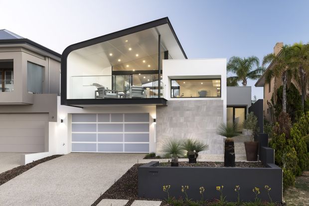 Contemporary multi-level residential facade with a dramatic cantilevered upper volume, curved dark metal edge, expansive glazing, recessed balcony, and bright integrated lighting. The lower level features a wide translucent-panel garage door, pale textured masonry cladding, a projecting white rectangular volume, and a landscaped front garden with sculptural planters and tropical planting.