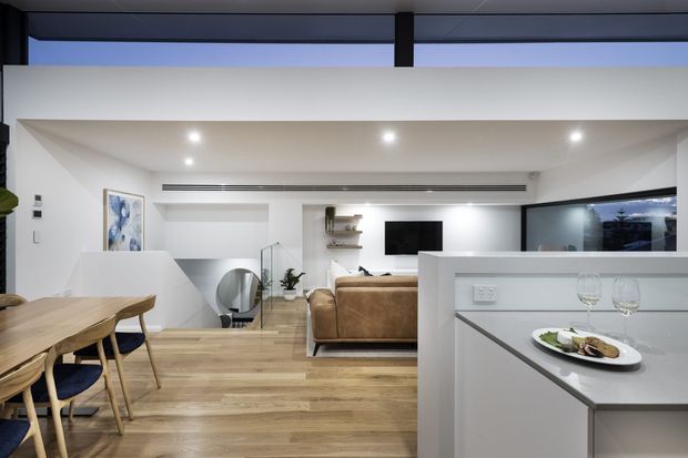 Open-plan contemporary apartment interior showing a kitchen island in the foreground, dining area to the left, and living room beyond. The space has light timber flooring, white walls and cabinetry, recessed downlights, integrated linear air conditioning, large dark-framed glazing, and a compact staircase with glass balustrade.