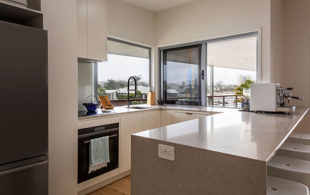 Bright contemporary kitchen with white full-height cabinetry, integrated appliances, a large pale stone island benchtop, and expansive corner glazing opening toward an outdoor terrace. The kitchen has a clean, refined fit-out with timber flooring and abundant natural light.