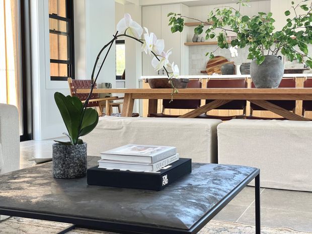 Bright open-plan living and dining area with a low, dark coffee table in the foreground, cream sofas, a timber dining table with woven chairs, and abundant indoor plants. The background includes white built-in cabinetry and open timber shelving, creating a warm, refined contemporary interior.