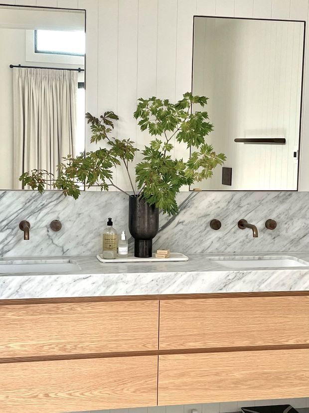 A refined bathroom vanity with a wide double basin, white veined marble wall and countertop surfaces, warm timber drawer cabinetry, dark bronze wall-mounted tapware, and a large mirror. A sculptural vase with leafy greenery sits centrally on the vanity, with a curtained window reflected in the mirror.