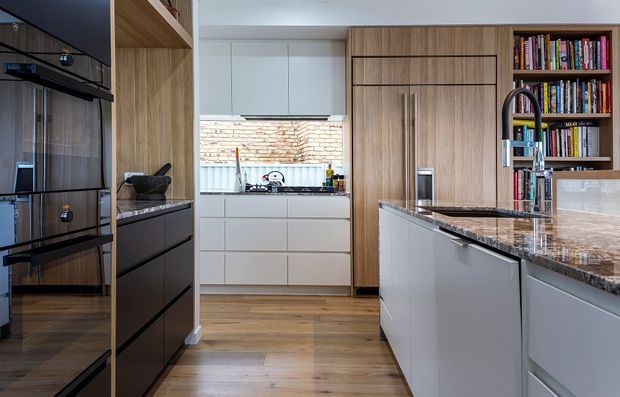 A contemporary galley-style kitchen viewed toward a rear preparation area. The space features full-height timber-look cabinetry, white handleless cabinetry, integrated black appliances, a dark polished stone island benchtop with waterfall edge, timber flooring, open shelving with books, and a large black gooseneck tap. A brick wall is visible through the rear window.