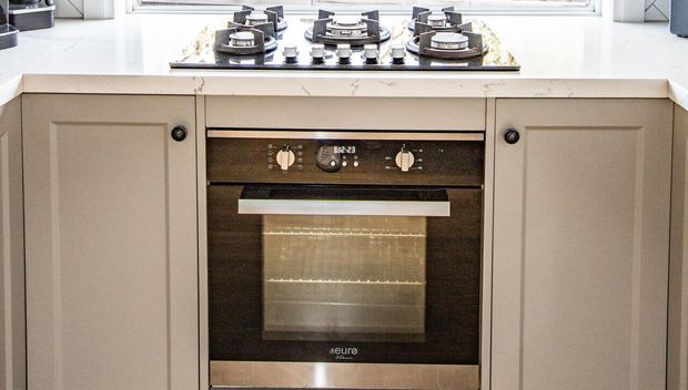 Close-up view of a fitted kitchen cooking area with a four-burner gas cooktop set into a pale stone benchtop and a built-in stainless-steel oven below. Taupe-grey shaker-style cabinetry with dark round knobs surrounds the appliance, with a bright window above.