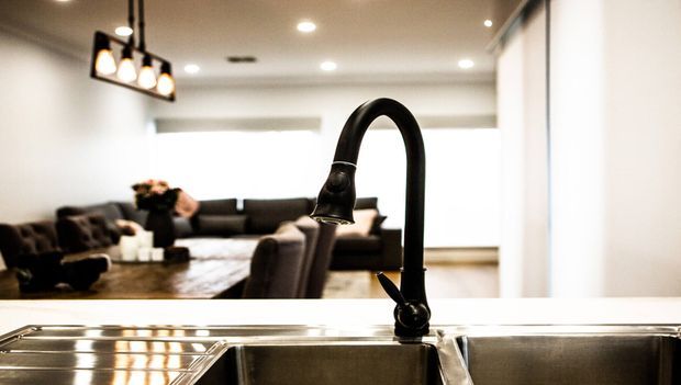 A close-up view of a black gooseneck kitchen faucet and stainless-steel double sink, with an open-plan living room visible beyond. The interior has white walls, recessed ceiling lights, dark upholstered seating, and a neutral contemporary fit-out.