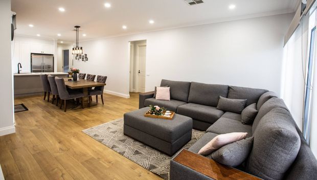 A bright open-plan living and dining area with a large dark grey sectional sofa and ottoman in the foreground, a timber dining table with upholstered chairs behind it, and a compact kitchen visible at the rear. The room has white walls and ceiling, warm timber-look flooring, recessed downlights, pendant lighting over the dining table, and large windows along the right side.