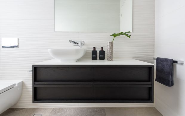 Contemporary bathroom vanity with a wall-mounted dark timber-look cabinet, white vessel basin, wall-mounted chrome tapware, large frameless mirror, textured light wall tiles, and a pale stone-look floor. A towel rail and neatly styled accessories complete the clean, refined composition.