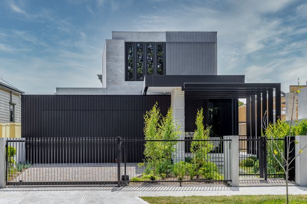 Street-facing view of a contemporary multi-level residence with a strong geometric composition. The facade combines dark vertical screening, pale rendered or concrete-like wall planes, narrow upper windows, and a projecting flat-roofed entry canopy with black framing. A gated driveway, front garden, fencing, and neighbouring homes are visible.