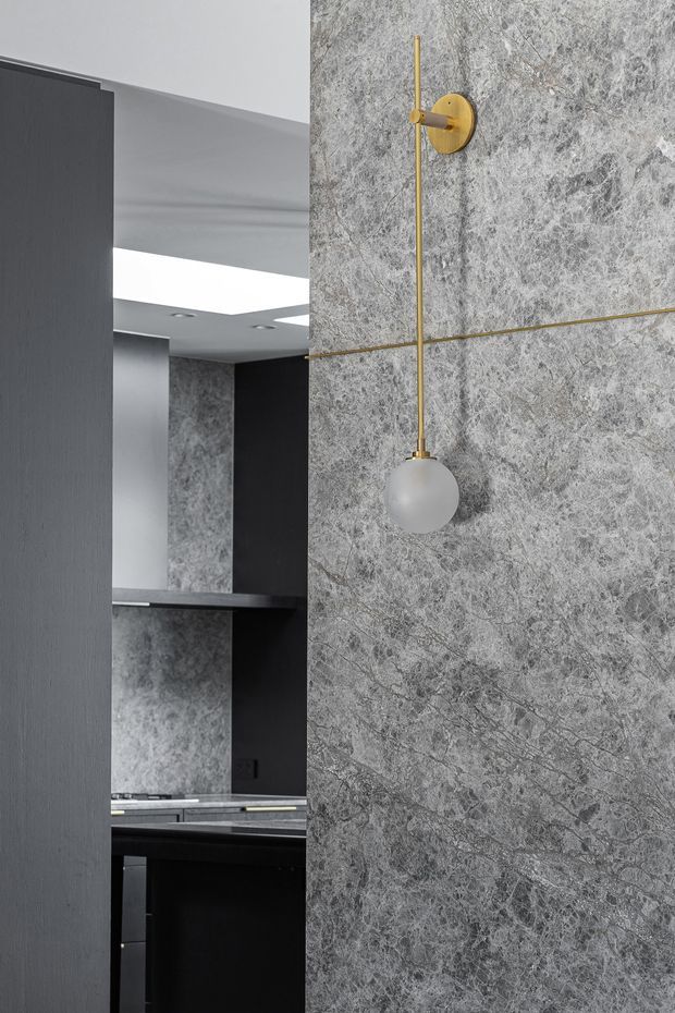 A close-up architectural interior view showing a sophisticated grey stone-clad feature wall with a minimalist brass wall light and opal globe shade. A dark, highly refined kitchen with black cabinetry and grey stone surfaces is partially visible through the opening to the left. The space has strong geometric lines, recessed ceiling lighting, and a restrained monochromatic palette with brass accents.