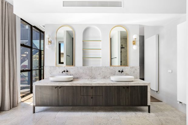A bright, refined double vanity bathroom with a long dark timber floating cabinet, pale stone benchtop, twin vessel basins, arched mirrors with brass trim, integrated open shelving, and decorative wall sconces. Large black-framed glazing and curtains appear to the left, while a shower area is visible in the mirror reflection.