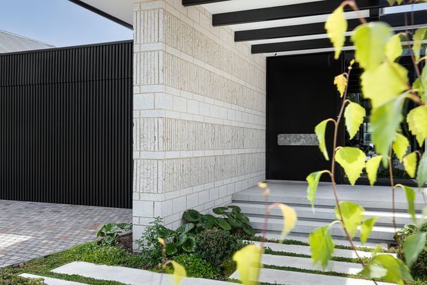 Contemporary residential entry facade with a pale textured masonry wall, black vertical slatted screening, a dark pivot entry door, and a covered porch framed by exposed black beams. Broad pale steps and rectangular stepping slabs lead through a planted garden bed to the entrance.
