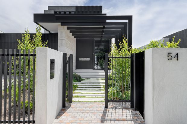 Front exterior view of a contemporary residence with a bold dark projecting entry canopy and repeated overhead beams, textured light-rendered walls, black vertical metal fencing, landscaped entry planting, and a paved path leading to the recessed front door. The house number 54 is displayed on the right boundary wall.