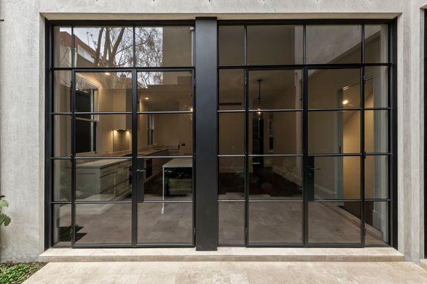 A contemporary black-framed steel-look glazed facade viewed from outside, with expansive gridded windows and a central dark mullion. Through the glazing, a refined interior is visible with warm lighting, a kitchen or kitchenette, polished neutral flooring, and a dark seating area. The exterior wall is light textured plaster or render, and the scene appears to open onto a paved terrace or courtyard.